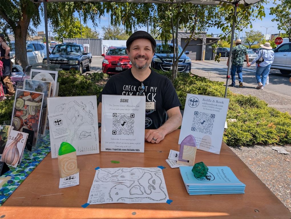 The author at Santa Rosa Zine Fest 2025. The table in front of the author has various games, signs, and other products the author has made on it.